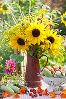 Bouquet of sunflowers and perennials Persicaria, Verbena bonariensis and Solidago in enamel jug with harvested vegetables in summer garden.