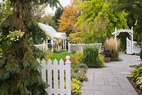 Grey flagstone path leading to a arbour through a white picket fence in front garden in autumn. Plants include Sedum spurium, Alchemilla mollis, Kleine 'Silberspinne', Miscanthus sinensis and a Robinia pseudoacacia 'Frisia' tree in the background. Il Etait Une Fois garden, Monteregie, Quebec, Canada. 
