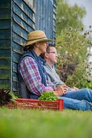Man and woman sitting down against an allotment shed, crate of harvested produce in the foreground