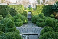 A formal garden featuring masses of clipped box topiary framed by yew hedges, paving that combines paving slabs and smaller setts to create decorative interest, and steps leading up to a kitchen garden dominated by four standard fig trees. 
