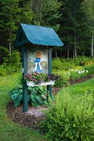 Spiraea x bumalda 'Goldmound' - Spirea shrub, Hosta - 'Blue Blazes' at the base of a wooden arbour decorated with an acrylic painting of a young woman gardener and a box of planted pink Impatiens in backyard Country garden in summer,  Jardin des Mesanges garden, Quebec, Canada
