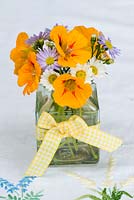 A summer posie of everyday garden flowers with Nasturtium, Aster and Anthemis in a glass jar decorated with yellow gingham ribbon.