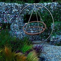 Modern, steel swing seat. Boundary wall of flints in wire gabions. Pebble floor runs between beds of low maintenance, drought resistant ornamental grasses. 