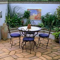 Moroccan style courtyard. Terracotta tiles, wire chairs and tiled table. Mosaic on wall above pool fed by gushing pitcher. Raised beds of arum, phormium, fatsia and grasses.