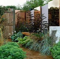 Planked path and wooden seat edged in hosta, black elder, Verbena bonariensis, achillea and grasses. Behind, stainless steel panels coated in titanium.