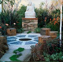 Tranquil meditation garden. Six oak cube seats set around circular pebble mosaic - wheel of life. Orange and maroon plants: kniphofia, heuchera, elder, sedum, sedge. Buddha.