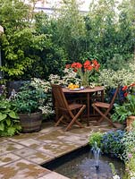 Edged in foliage, table laid with fruit and vase of gerbera. Paved terrace, sunken pool with fountain. Planting - dogwood, hydrangea, ceanothus, diascia, alyssum.