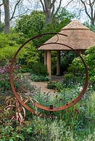 A contemporary cottage garden with circular metal sculpture and thatched gazebo.