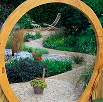 Seen through moongate, feng shui garden of rocks, grasses and flowers with winding brick and gravel path joining different areas.