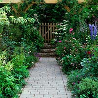 In front garden, wood gate opens onto stone path edged in roses, delphinium, hardy geranium, cistus, salvia and astrantia.