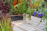 A seating area beside a water tank, with planting of Imperata cylindrica 'Rubra', Campanula, Cosmos atrosanguineus, Ajuga reptans 'Braunherz' and Anthemis tinctoria 'Sauce Hollandaise' beside a water tank and metal grid path. Garden - A Space to Connect and Grow.