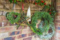 Mixed evergreen wreath hanging on a rustic tool rack, with a helichrysum italicum wreath and mixed evergreen hearts. Foliage includes Sequoiadendron giganteum, Pinus, Larch and Ilex aquifolium