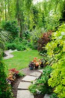 Neatly kept garden with stepping stones, lawn, mixed shrubs, birch trees and pots with hostas and heucheras