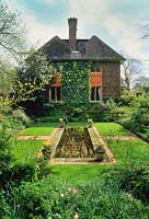 Formal sunken garden with rectangular pond set within lawn. Stone and brick detailing. Neatly clipped panel of ivy on house wall.