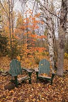 Two old green painted wooden adirondack chairs in a deciduous tree forest of Acer and Betula in backyard country garden.  Laurentians, Quebec, Canada