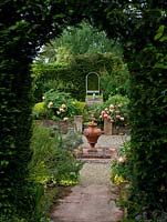 View across formal, sunken garden edged in raised beds of shrubs, irises, perennials and peonies - Paeonia x lemoinei 'Chromatella' and 'Souvenir de Maxime Cornu'. Central urn.