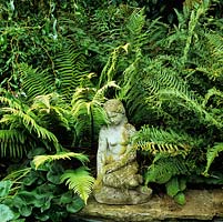 Beside pond, stone statue of girl rests in shade of ferns and contorted willow.