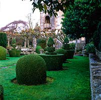Topiary shapes in clipped box march across lawn leading to Arts and Crafts house flanked by stone troughs and avenue of limes.