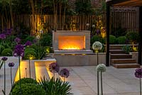 Town garden designed by Kate Gould, lit at night. Flames from an open gas fireplace illuminate the sunken terrace which is edged in beds of box balls interspersed with purple and white allium. Back boundary is planted with tall golden bamboo.