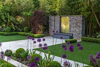 Town garden designed by Kate Gould. A stainless steel water feature is set into a dry stone wall. Rectangular patches of lawn are edged in box balls interplanted with purple and white allium. Boundary beds are filled with bamboo, cordyline, acer, Trachycarpus fortunei, hosta and euphorbia.