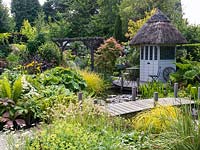 A bridge crosses a lush bog garden and natural pond. Planting includes Ligularia dentata, Gunnera manicata, Rodgersia, Rheum palmatum, Trollius x cultorum. and Carex oshimensis Evergold.