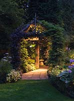 Garden lit at night. Wooden pergola clad in vine, clematis and wisteria leads to staddle stone. In beds, hosta, phlox, hardy geranium and box.