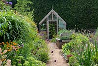 A path leads to a greenhouse through a double border planted with late summer flowering perennials including echinops, cardoon, salvia, daylily, crocosmia, sedum, fennel, helenium, Verbena bonariensis, Carthusian dianthus, and drumstick allium. On right, raised bed planted with herbs.