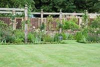 View across lawn to pergola with lamps, pyramids of clipped Buxus sempervirens, Rosa 'Mutabilis', Iris and Penstemon - Priory House, Wiltshire