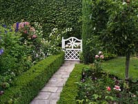 White gate opens onto stone path between box-edged beds of apples, clematis, delphinium, hardy geranium, philadelphus and roses, offset against tall beech hedge.