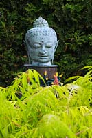 Close-up of buddha head sculpture and Rhus typhina 'Tiger Eyes' - Sumac shrub in Japanese garden in private backyard garden in summer