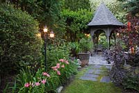 Illuminated khaki painted wooden lattice gazebo on paving stone patio and pink Lilium 'Rodeo' - Lilies, Prunus cistena 'Purpleleaf Sand Cherry'. Planter with Cyperus papyrus 'King Tut' - Egyptian Paper Rush in private backyard garden in summer