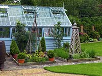 A vegetable garden with small raised beds in front of a painted wooden framed greenhouse.
