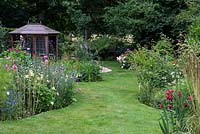 A grass path between a colourful wildflower border and a mixed herbaceous border, with a wooden summerhouse behind.