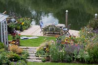 Bird's eye view of summerhouse by bed of catmint, coneflower, annual poppies and oriental lilies. Lawn leads to riverside deck edged in pots and beds of oriental lilies, daylilies, Verbena bonariensis, annual poppies, agapanthus and kniphofia.