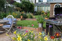 Riverside deck with barbecue, dining table and chairs, is edged in pots and bed of oriental lilies, daylilies, Verbena bonariensis and annual poppies. A step up leads to a lawn and raised bed.