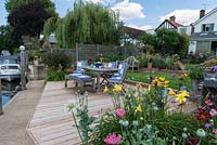 Riverside deck with dining table and chairs edged in pots and beds of oriental lilies, daylilies, Verbena bonariensis, annual poppies, agapanthus, dogwood and kniphofia.
