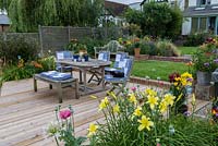 Riverside deck with dining table and chairs, is edged in pots and beds of oriental lilies, daylilies, Verbena bonariensis, annual poppies, agapanthus, dogwood and kniphofia.