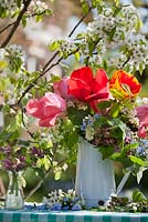 Display of spring flowers in enamel jug, includes tulips, Prunus padus - bird cherry, Myosotis arvensis, Allaria petriolata - garlic mustard, Lamium orvala. Flowering pear tree behind - Pyrus 'Williams'.