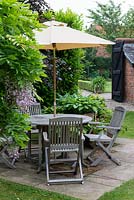 Wooden garden furniture on a small stone patio in front of a small container garden planted with Hosta 'Great Expectations', Hosta 'June' and at back Hosta 'Silk Road'.