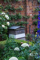 A beehive in front of an apple espalier trained against a brick wall.