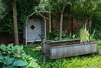 A shady corner of a walled garden with covered wooden seat, a small pond made from a galvanised metal animal feeding trough and shade tolerant planting including hosta and box.