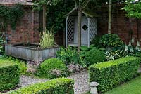 A shady corner of a walled garden with covered wooden seat, a small pond made from a galvanised metal animal feeding trough and shade tolerant planting including hosta and box.