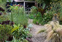 A gravel garden with Stipa grasses, stone statue and borders with tropical foliage plants and perennials. The path leads to a covered seating area.