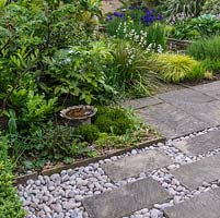Detail of paving slabs interspersed with grey pebbles. Small bird bath in border, edged in hebe, fern and fatsia. Behind, white sisyrinchium, blue Siberian iris, Hakonechloa macra, hosta and pink bistort.