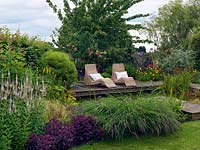 Wicker loungers on the pond side deck, behind a border including Veronicastrum virginicum 'Fascination',  Sedum telephium 'Purple Emperor' and Pennisetum alopecuroides,