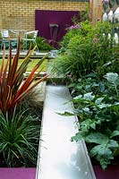 Purple wall with water spout and stainless steel rill surrounded by Phormium - New Zealand flax, Acanthus - Bears breech, Stipa - Spear grass, and Verbena. Designed by Paul Dracott.