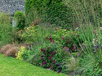 A traditional long mixed border with roses, Stipa and Carex grasses, and herbaceous plants including Astrantia 'Roma', Euphorbia 'Fireglow' and Alchemilla mollis