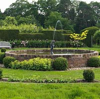 Formal raised circular pool with fountain, edged in box balls and Alchemilla mollis. Behind, peony cutting bed against old yew hedge.