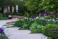 The Time In Between by Husqvarna and Gardena. View of path made from natural sandstone paving slabs separated with Pratia pedunculata and surrounded by Zantedeschia aethiopica, Allium giganteum, Alchemilla mollis, Pogonatherum paniceum, Orlaya grandiflora 