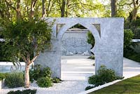 view of a seating area with cushions against marble wall with Arabic writing through an arch surrounded by Punica granatum, Cistus salviifolius, Alternanthera green. The Beauty of Islam - Chelsea Flower Show 2015